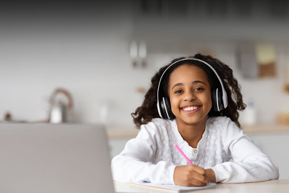 Young girl in a white sweater holding a notebook and pencil and looking at the camera smiling while taking a class at Alabama Connections Academy.