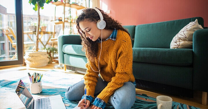 Online high school student sitting on the floor in front of a laptop and writing in a notebook