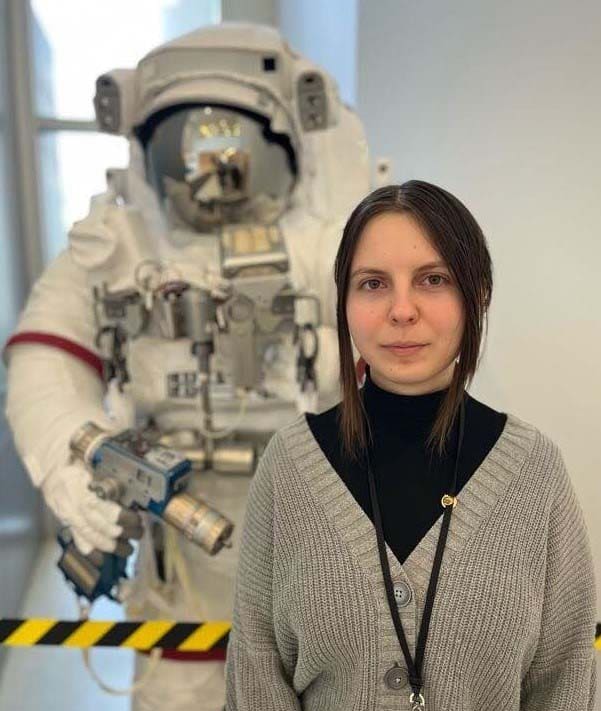Eva T. from Ohio Connections Academy in a black turtleneck shirt and brown sweater standing in front of a space suit display at NASA Glenn Research in Cleveland. 
