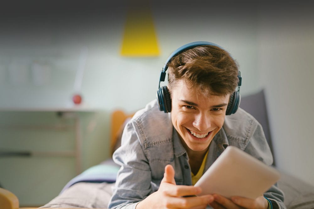 Young teen boy wearing a blue shirt and headphones and looking at a tablet while taking an online class at Wyoming Connections Academy.