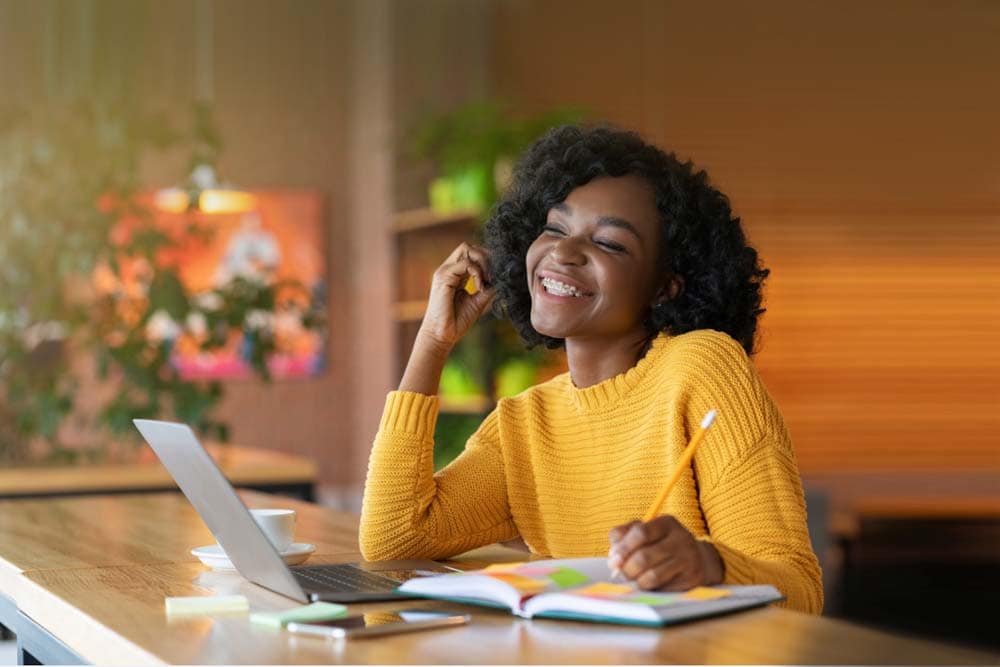 Young girl in yellow holding a notebook and pencil and looking at a laptop