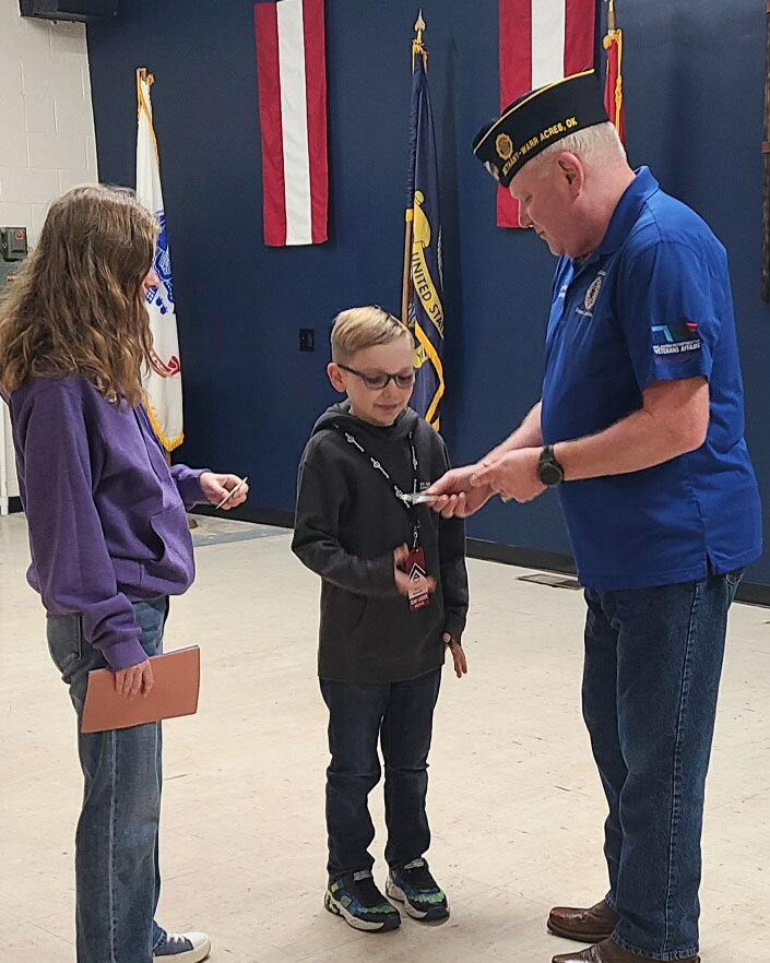 A man in a blue uniform and hat hands an item to a young boy, while a girl in a purple sweatshirt stands nearby holding a book. They are indoors with flags in the background.