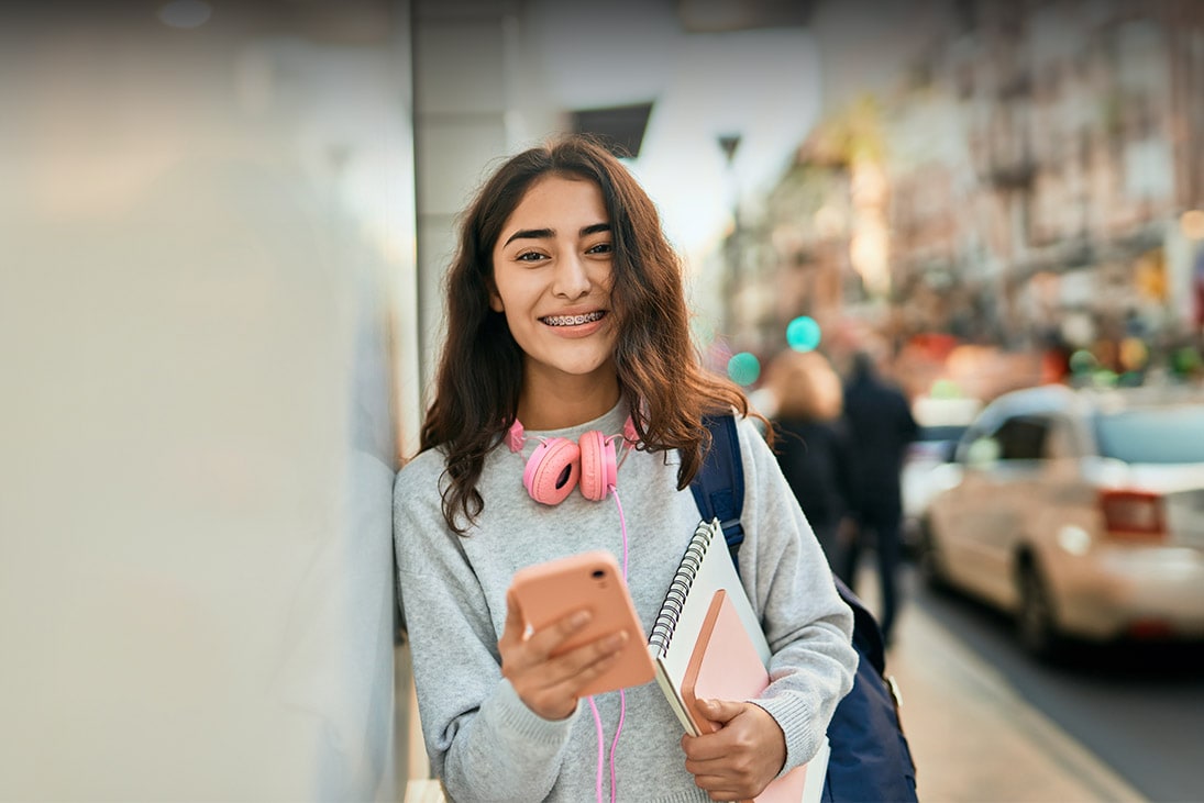 An online school student leaning against a wall and holding a pink phone and a notepad